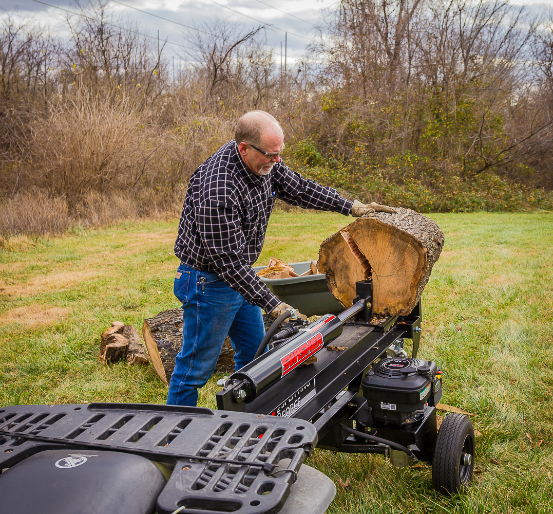 A yellow log splitter cutting a short log in half. The splitter is pushed up against a large pile of small logs.