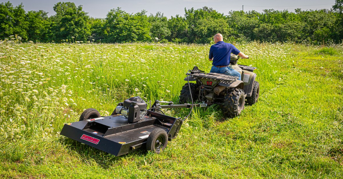 A man rides an ATV through a grassy field while towing a brush mower attachment, cutting a path through tall grass.