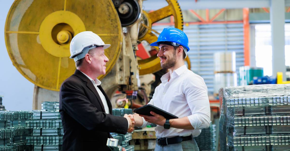 Two men in hard hats shaking hands. They are standing in an industrial facility with stacks of metal components around them.