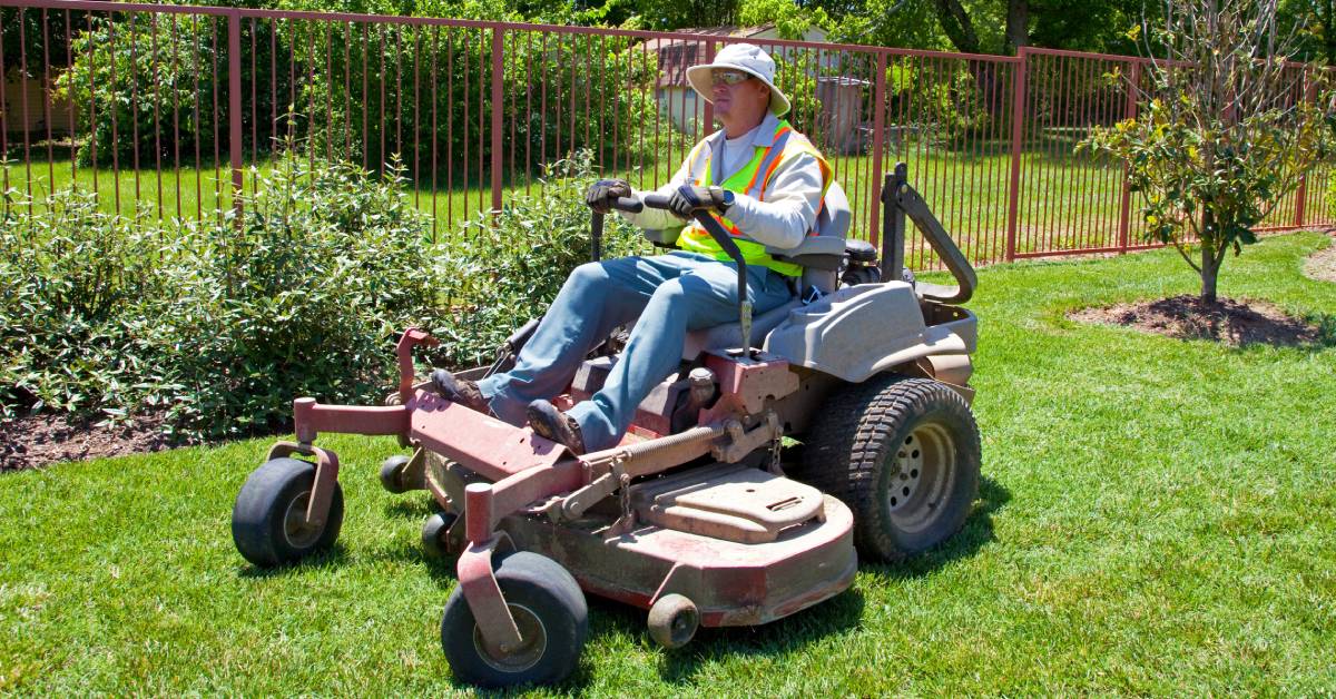 An older man wearing a reflective vest riding a zero-turn lawn mower. He is mowing the lawn of a commercial property.