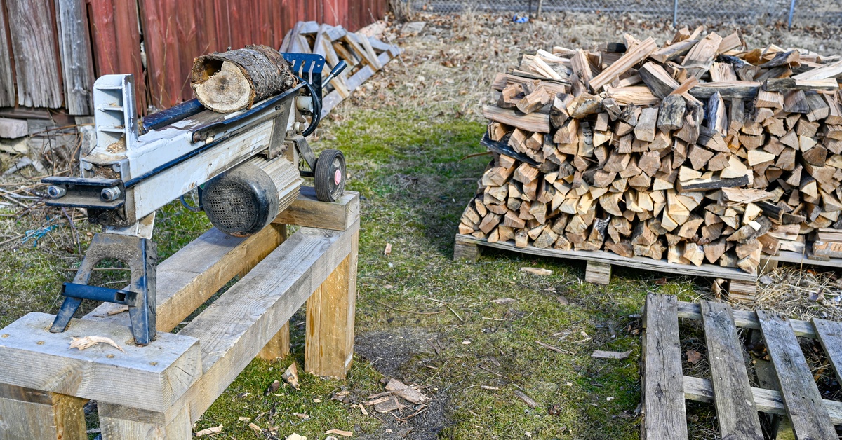 An electric log splitter sitting on top of a wood surface. There is a large pile of chopped wood in the background.