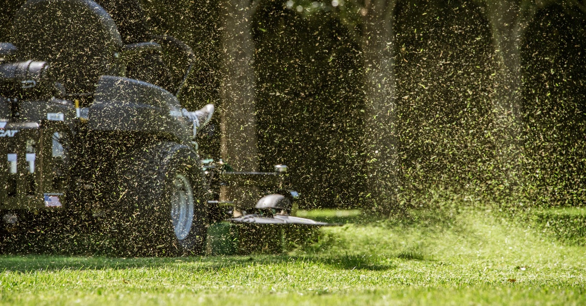 A person driving their zero-turn mower past a row of trees while hundreds of blades of grass fly through the air.