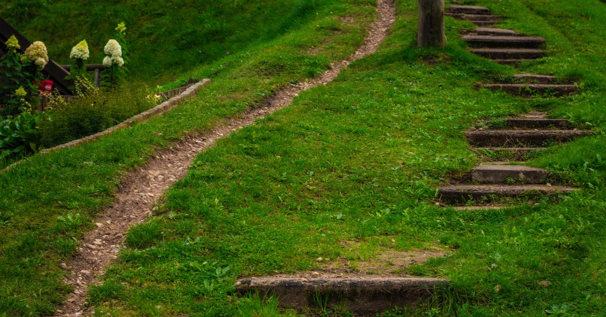 A grassy trail leading up a hill that has a worn path and some dirt steps. There is a small garden near the trail.